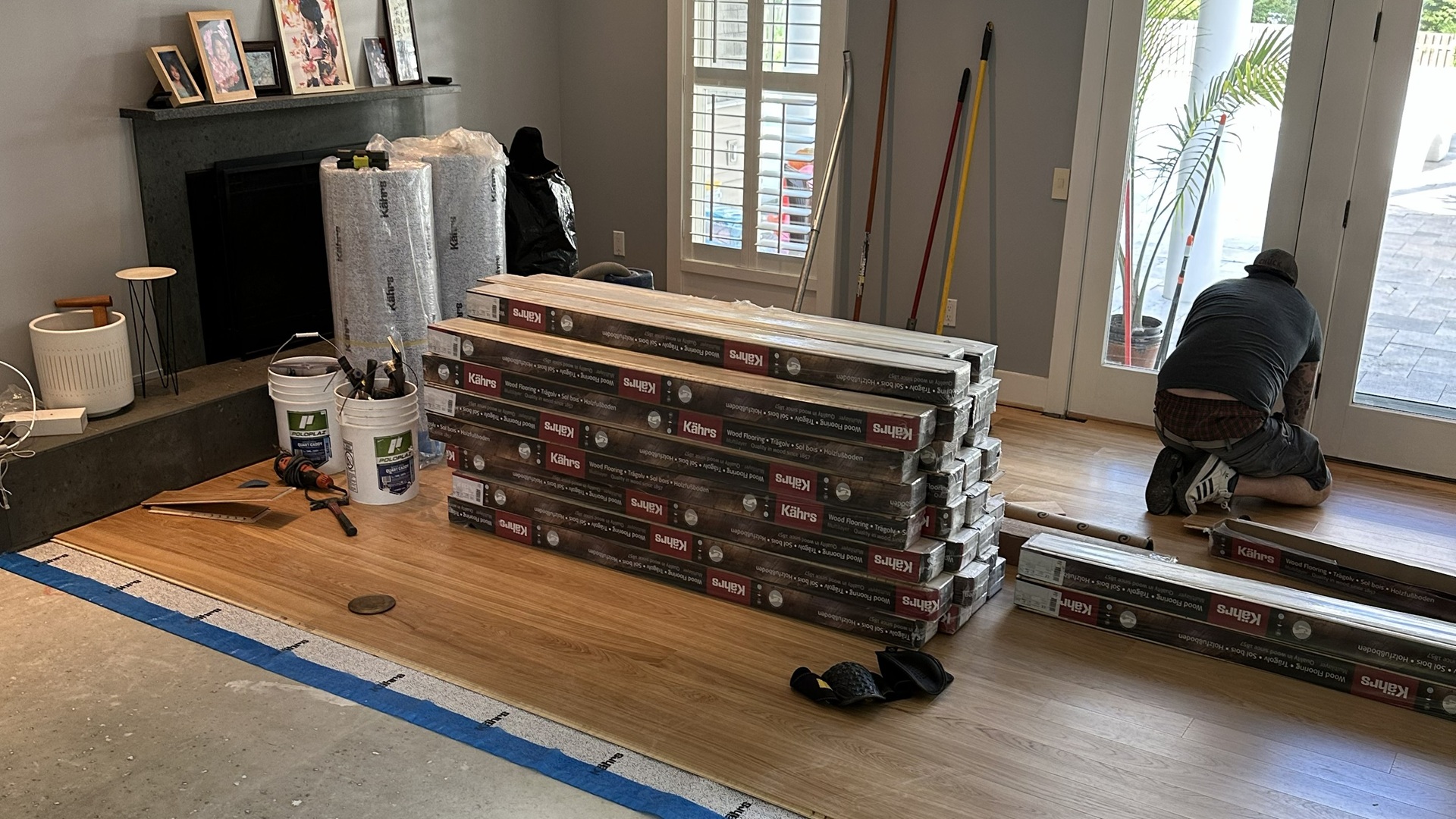 A professional installer laying luxury vinyl plank (LVP) flooring in a living room next to a stack of flooring materials and an exposed subfloor.